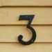 Close-up of a black number three sign mounted on a textured yellow wooden wall.